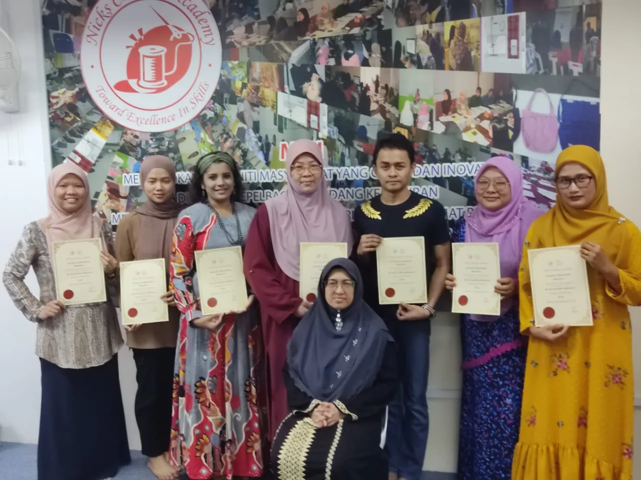 A group of graduates holding their certificates in front of the academy's logo wall